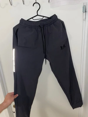 Svarta mjukisbyxor  Under Armour - Svarta mjukisbyxor från Under Armour i storlek M. Byxorna har elastisk midja med snörning, två sidofickor och en reflekterande logga längs benet. Materialet är mjukt och lätt polyester, perfekt för träning eller chill. Loose fit och smalare benslut.Liknande modell som originala.