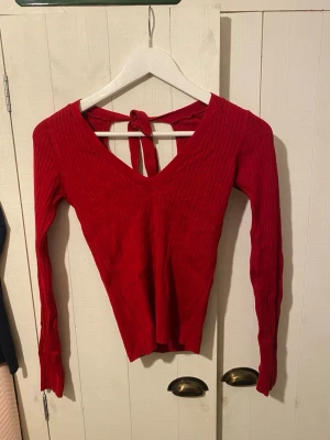 Röd ribbad topp från Stradivarius - Säljer en snygg röd långärmad topp från Stradivarius i storlek S. Toppen har ribbad struktur, v-ringning och en öppen rygg med knytband. Perfekt för dig som gillar detaljer och vill ha en stilren men ändå unik look.