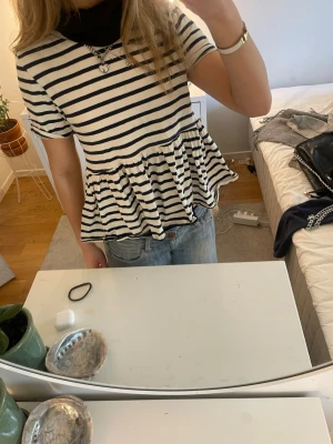 Randig volangblus från Zara - Säljer en söt blus från Zara i storlek S med marinblå och vita ränder. Toppen har korta ärmar, rund hals och en volang nedtill som ger en skön och avslappnad vibe. Materialet är mjuk bomull, perfekt för vår och sommar.