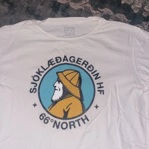 Vit t-shirt med tryck från 66°North - Säljer en vit t-shirt från 66°North i storlek S med ett stort grafiskt tryck på bröstet som visar en profil av en fiskare i gul regnjacka och hatt. Rund hals och korta ärmar. Perfekt för dig som gillar coola prints och islandsvibbar.