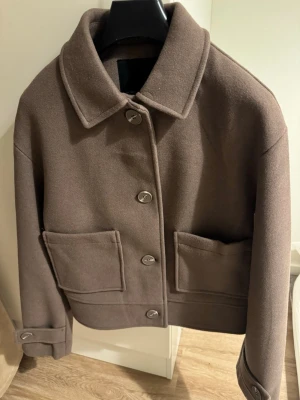 Brun ulljacka med stora fickor - Snygg brun jacka i ull med klassisk krage och stora framfickor. Jackan har knappar hela vägen och detaljer vid ärmsluten. Modellen är boxig och lite kortare, perfekt för dig som gillar en clean och chill look.