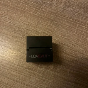 Huda Beauty setting powder - Säljer ett Huda Beauty löspuder i en svart fyrkantig förpackning med transparent botten. Pudret är ljusbeige och kommer med en rund applikator för enkel applicering. Perfekt för att fixera smink och ge en matt finish. Den är i färgen Banana Bread och den är använd