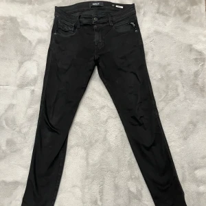 Svarta Replay Anbass jeans - Snygga svarta jeans från Replay, modell Anbass. De har en slim passform med klassisk femficksdesign och diskreta detaljer på bakfickorna. Jeansen är tillverkade i stretchigt bomullsmaterial för extra komfort och har en modern look som passar till det mesta.