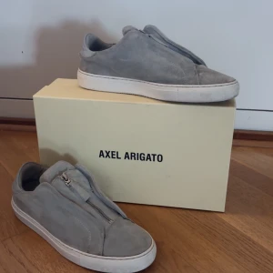 Grå mockasneakers från Axel Arigato - Stilrena sneakers i ljusgrå mocka från Axel Arigato med vit platt sula och dragkedja framtill istället för klassisk snörning. Snygg minimalistisk design. Använda ett tag men fortfarande i bra skick!