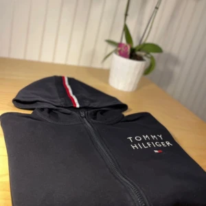 Svart hoodie Tommy Hilfiger med dragkedja - Svart hoodie från Tommy Hilfiger med hel dragkedja, huva och klassisk logga på bröstet. Snygg detalj med röd, vit och marinblå rand längs huvan. Mjuk bomullskänsla och ribbade muddar. Perfekt för en chill och stilren look.