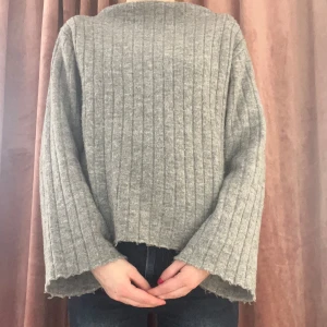 Grå ribbad stickad tröja - Mysig grå stickad tröja med ribbat mönster och vida ärmar. Tröjan har en avslappnad passform och råa kanter vid ärmslut och nederkant. Perfekt för lager-på-lager och chill dagar.