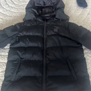 Svart pufferjacka Urban Classics - Svart pufferjacka från Urban Classics med huva och dragkedja framtill. Jackan är vadderad och har två snedställda fickor. Perfekt för kalla dagar och har en clean, stilren look som funkar till det mesta. Riktigt skön och bästa jackan till vinter som håller dig varm.