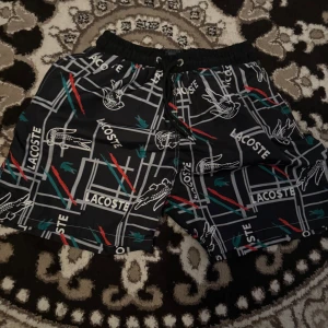 Svarta lacoste badbyxor - Snygga svarta shorts från Lacoste med grafiskt mönster, vita och röda linjer samt gröna detaljer. Shortsens design har Lacoste-loggan och krokodiler över hela tyget. Elastisk midja med snörning för skön passform. Perfekta för chill dagar eller häng med kompisar.