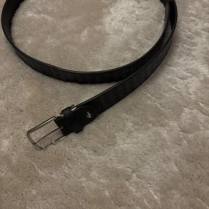 Lacost bälte  - Snyggt svart bälte från Lacoste i äkta skinn med diskret geometriskt mönster och silverfärgat spänne. Klassisk design med Lacoste-logga vid spännet och stilrena detaljer. Perfekt accessoar för att lyfta din outfit.Bältet är helt nytt och inte använt köpt men inte kommit till nytta. Ny pris ligger på 800kr. 