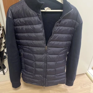 Mörkblå dunjacka från Moncler - Snygg svart dunjacka från Moncler med quiltad front och stickade ärmar. Jackan har dragkedja framtill, Moncler-logga på ärmen och ribbad krage. Perfekt för dig som vill ha en stilren och bekväm jacka med exklusiv känsla.