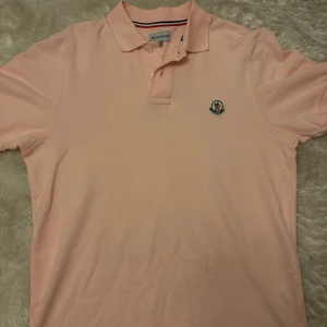 Ljusrosa Moncler piké t-shirt M - Snygg ljusrosa piké t-shirt från Moncler i bomull. Klassisk krage med två knappar och broderad Moncler-logga på bröstet. Kortärmad och med stilrena ränder i kragen. Perfekt för dig som gillar clean och exklusiv vibe. Den är endast använd 1 gång och bara ligger nu. HÖR AV VID FRÅGOR!