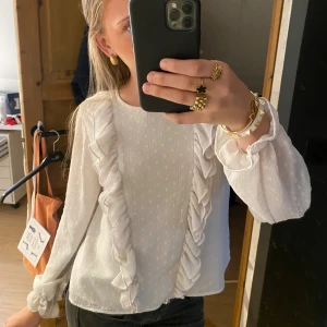 Vit blus med volangdetaljer - Supersöt vit blus med diskret prickstruktur och volanger längs framsidan. Långärmad modell med lätt transparent tyg och knäppning bak i nacken. Säljer då den inte kommer till användning!🥰