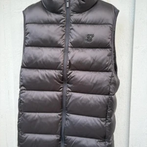 Svart dunväst från SikSilk - Säljer en svart dunväst från SikSilk med diskret logga på bröstet. Västen har quiltad design, hög krage och dragkedja framtill. Perfekt att slänga över en hoodie för extra värme. Stilren och enkel look som funkar till det mesta.