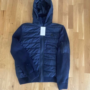 Mörkblå cardigan från Pier One - Snygg mörkblå cardigan från Pier One med huva och ribbade muddar. Jackan har quiltad framsida och rygg, stickade ärmar och dragkedja framtill. Perfekt för kyliga dagar och har en stilig look. Det står att det är en S men passar nåt större 