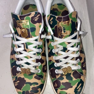 Adidas Stan Smith x Bape camo sneakers - Unika Adidas Stan Smith x Bape sneakers med ikoniskt grönt, brunt och beige camo-mönster. Skorna har vita snören, gulddetaljer och branding på häl och plös. Klassisk rund tå och platt sula, tillverkade i skinn. Perfekt för dig som vill sticka ut med streetwear.