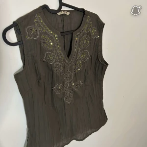 Broderad militärgrön blus från TYK - Supersnygg militärgrön blus från TYK med broderade detaljer och små paljetter framtill. Toppen är ärmlös med v-ringning och har ett lätt skrynklat tyg som ger en avslappnad vibe. Perfekt för dig som gillar unika detaljer och boho-stil.