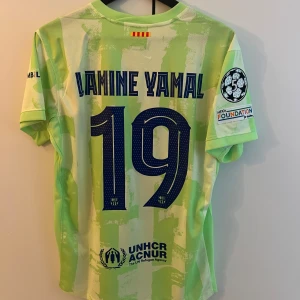 Barcelona Lamine Yamal matchtröja - Säljer en Barcelona fotbollströja med Lamine Yamal och nummer 19 på ryggen. Tröjan är ljusgrön med gröna mönster, har korta ärmar och är tillverkad i lätt polyester. FCB-emblem, Nike-logga och Spotify-sponsor på framsidan, samt UEFA Foundation-märke på ärmen. Replika