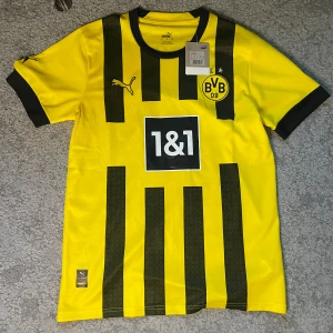 Borussia Dortmund fotbollströja - Borussia Dortmunds officiella hemmatröja från Puma med klassiska svarta ränder på gul botten. Tröjan har klubbmärke och sponsortryck på bröstet, svart krage och svarta muddar. Materialet är lätt och andas, perfekt för match eller träning.