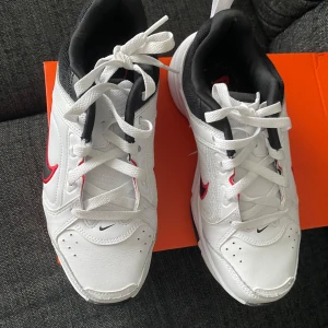 Vita Nike sneakers med röd Swoosh - Säljer ett par fräscha vita Nike sneakers med svart och röd Swoosh på sidorna. Skorna har snörning, svart insida och detaljer i syntet och skinn. Sulan är svart och vit med tydlig Nike-logga undertill. Perfekta för dig som gillar klassisk sportig stil.