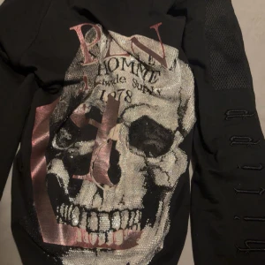 Svart  zip hoodie från Philipp Plein med dödskalle - Cool svart hoodie från Philipp Plein med stor dödskalle och glittriga detaljer framtill. Har patchar med dödskalle och logga på bröstet, samt dragkedja och huva. Snygga detaljer på ärmarna och en unik look som sticker ut. Väldig sällsyn tröja eftersom den är mer än 6 år gammal. STORLEK M MEN PASSAR L  