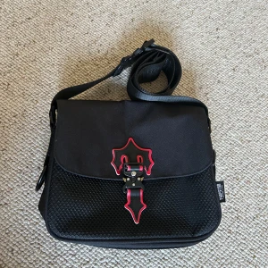 Svart axelväska från Trapstar - Säljer en svart side bag från Trapstar. Väskan har justerbar axelrem, röd dragkedja på baksidan och flera fack inuti. Den är i helt nytt skick. 