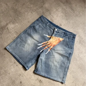 Blå jeansshorts med handtryck - Unika blå jeansshorts med coolt tryck av en hand med långa naglar över gylfen. Perfekt för dig som vill sticka ut med en edgy streetstil. Alla mått finns i bilden 