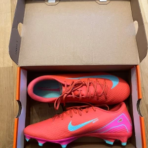 Nike Mercurial Vapor 16 fotbollsskor 42.5 - Nike Mercurial Vapor 16 Academy FG/MG i en riktigt snygg röd och turkos färg med rosa detaljer. Helt oandvända och perfekta för dig som vill sticka ut på fotbollsplanen, priset kommer ej att sänkas mer