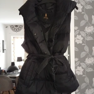 Svart dunväst från Pernilla Wahlgren - Snygg svart dunväst från Pernilla Wahlgren med huva och bälte i midjan. Västen har knappar framtill och en quiltad design som ger en cool och modern look. Perfekt att slänga över en hoodie eller tröja när det är kyligt ute.
