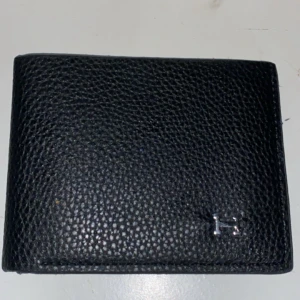 Svart plånbok i skinn - Säljer en stilren svart plånbok äkta skinn. Plånboken har flera kortfack och en klassisk H-detalj i metall på framsidan. Perfekt för dig som vill ha en lyxig och praktisk accessoar i fickan.