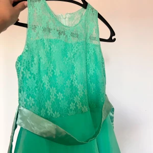 Grön spetsklänning med blommönster - Snygg grön klänning med ärmlös design och blommig spets upptill. Klänningen har ett satinband i midjan och en lätt, flowy kjol. Perfekt för dig som vill sticka ut med en fräsch färg och fina detaljer. Klänningen är lång ända ner till fötterna. 