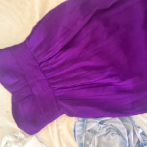 Lång lila klänning i chiffong - Säljer en elegant långklänning i stark lila färg med svepande, transparent chiffongtyg. Klänningen har axelbandslös design och ett vackert draperat liv som ger en snygg siluett. Perfekt för dig som vill sticka ut med färg och flowy vibes. Dock är den lite kronglig på dragkedjan men funkar. 