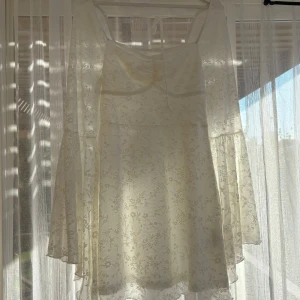 Vit blommig kortklänning med trumpetärm - Superfin vit kortklänning med diskret blommigt mönster och trumpetärmar. Klänningen har fyrkantig urringning och knyt i ryggen, vilket ger en romantisk vibe. Lätt och luftig, perfekt för varma dagar. Materialet känns som tunn bomull eller polyester.