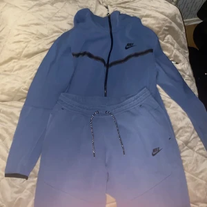 Nike Tech Fleece Tracksuit byxor & hoodie (300 st) - Säljer en blå hoodie från Nike med dragkedja och huva. Tröjan har svarta detaljer och Nike-logga på bröstet. Perfekt för en avslappnad stil. Byxorna är i ett bra skick hoodien är i ett okej skick, samt att byxorna är i storlek L och hoodien är i storlek M. Lite små hål i den 