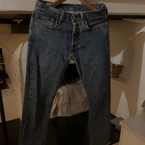 Levi's 501 blå jeans W28 L30 - Klassiska Levi's 501 jeans i blå denim med raka ben och femficksmodell. Jeansen har knappgylf, bruna sömmar och den ikoniska läderpatchen bak i midjan. Perfekta för dig som gillar tidlös stil och vill ha ett par riktigt schyssta jeans.