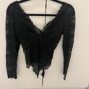 Svart spetsig långärmad body - Säljer en svart body i genombruten spets med v-ringning och långa ärmar. Toppen har fina spetsdetaljer längs kanterna och en snörning framtill som ger en extra snygg touch. Perfekt för dig som vill sticka ut lite extra.