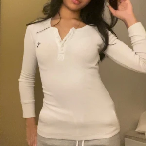 Vit långärmad tröja - Superskön vit tröja med lång ärm och knappslå framtill. Snyggt broderad logga på bröstet och figurnära passform. Perfekt till jeans eller mjukisbyxor för en chill look.