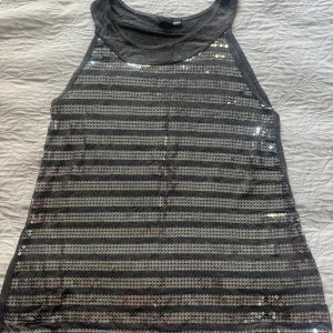 Glittrig randig linnetopp från H&M - Snygg grå linnetopp från H&M med silvriga paljetter i horisontella ränder på framsidan. Toppen är ärmlös och har en enkel, rak passform. Perfekt för dig som vill sticka ut med lite extra glitter och coola detaljer. Storlek och material lappar avklippt. 