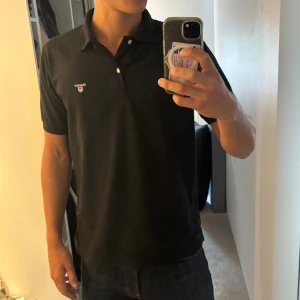 Svart pikétröja från GANT - Klassisk svart pikétröja från GANT med liten broderad logga på bröstet. Tröjan har krage, två knappar och korta ärmar. Perfekt till jeans eller shorts för en clean och stilren look.