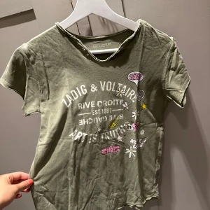 Grön Zadig & Voltaire t-shirt med tryck - Cool grön t-shirt från Zadig & Voltaire med vit text och färgglada tryck som blommor, stjärnor och peace-märke. Kortärmad modell i mjuk bomull med råa kanter vid halsen för en avslappnad vibe.