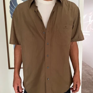 Brun kortärmad skjorta Sand - Snygg brun skjorta från sand  med korta ärmar och klassisk krage. Skjortan har en bröstficka med diskret logga och knäppning framtill med mörka knappar. Perfekt för en chill och stilren look. Unikt material av bomullsviskos.