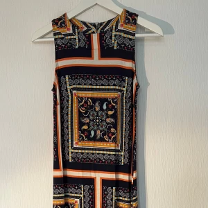 Mönstrad ärmlös klänning från Indiska - Säljer en cool ärmlös klänning från Indiska i storlek XS. Klänningen har ett färgstarkt paisley- och rutmönster i marinblått, orange, vitt och inslag av turkos. Den är rak i modellen och har en lång längd, perfekt för att sticka ut med sin unika design. Ny med prislapp! 