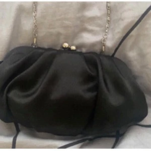 Svart kuvertväska med kedja - Snygg svart kuvertväska i satinliknande material med mjuka veck och guldfärgad kedja. Väskan har en klassisk metallknäppning upptill och passar perfekt när du vill lyfta din outfit med en elegant accessoar.