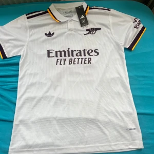 Arsenal vit fotbollströja Adidas M - Snygg Arsenal fotbollströja från Adidas i vit med lila och gula detaljer på krage och ärmslut. Tryck med 'Emirates Fly Better' på bröstet och Gyökeres 14 på ryggen. Klassisk pikékrage och korta ärmar. Tillverkad i lätt och ventilerande material.