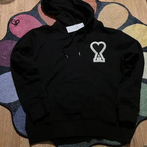 Svart hoodie från AMI med hjärtdetalj - Svart hoodie från AMI med stor vit broderad hjärtdetalj och bokstaven A på bröstet. Tröjan har huva med snörning, ribbade muddar och en klassisk känguruficka framtill. Perfekt för dig som gillar stilrena streetwear-plagg med unik design.