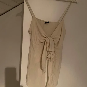 Beige linnetopp med knyt framtill - Snygg beige linnetopp med smala axelband och en stor knytning framtill. Toppen har en enkel och stilren design som passar perfekt till sommaren. Lätt och luftig känsla, perfekt för varma dagar.