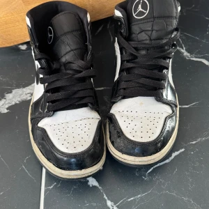 Nike Air Jordan 1 svartvita sneakers - Svartvita Nike Air Jordan 1 sneakers med klassisk hög siluett och ikoniska vingar-loggan på sidan. Skorna har svart snörning, vit tåbox med perforeringar och svart sula. Materialet är en mix av skinn och syntet. Perfekta för dig som gillar streetstyle och vill sticka ut.