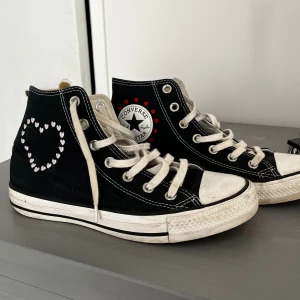 Converse All Star svart med hjärtan - Svarta Converse All Star high-tops med vita snören och sula. Skorna har broderade hjärtan i vitt och rött på sidorna och runt loggan. Klassisk rund tå och platt sula. Perfekt för dig som vill ha en personlig touch på dina sneakers💕💕 Priset kan diskuteras vid snabb affär!! 💕