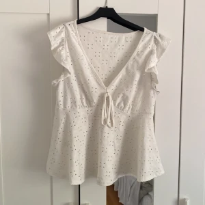 Vit blus med volang och knyt - Supersöt vit blus i bomull med broderade hål, volangdetaljer på axlarna och v-ringning med knyt framtill. Ärmlös och lätt utsvängd passform.