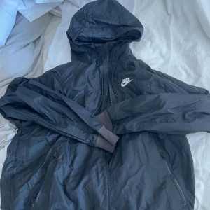 Svart vindjacka från Nike med huva - Svart vindjacka från Nike med huva och dragkedja framtill. Jackan har långa ärmar med muddar och två fickor med dragkedja. Perfekt för blåsiga dagar och enkel att slänga på sig när du är på språng. storlek S, ny kostar 1000kr frågor? skriv. 