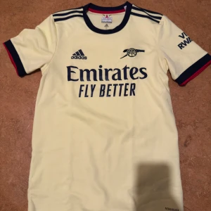 Arsenal bortatröja Adidas gul S - Arsenal bortatröja från Adidas i ljusgul färg med mörkblå detaljer och broderad logga. Tröjan har korta ärmar, rund hals och tryckta sponsorloggor. Tillverkad i funktionsmaterial som andas, perfekt för fotboll eller träning.